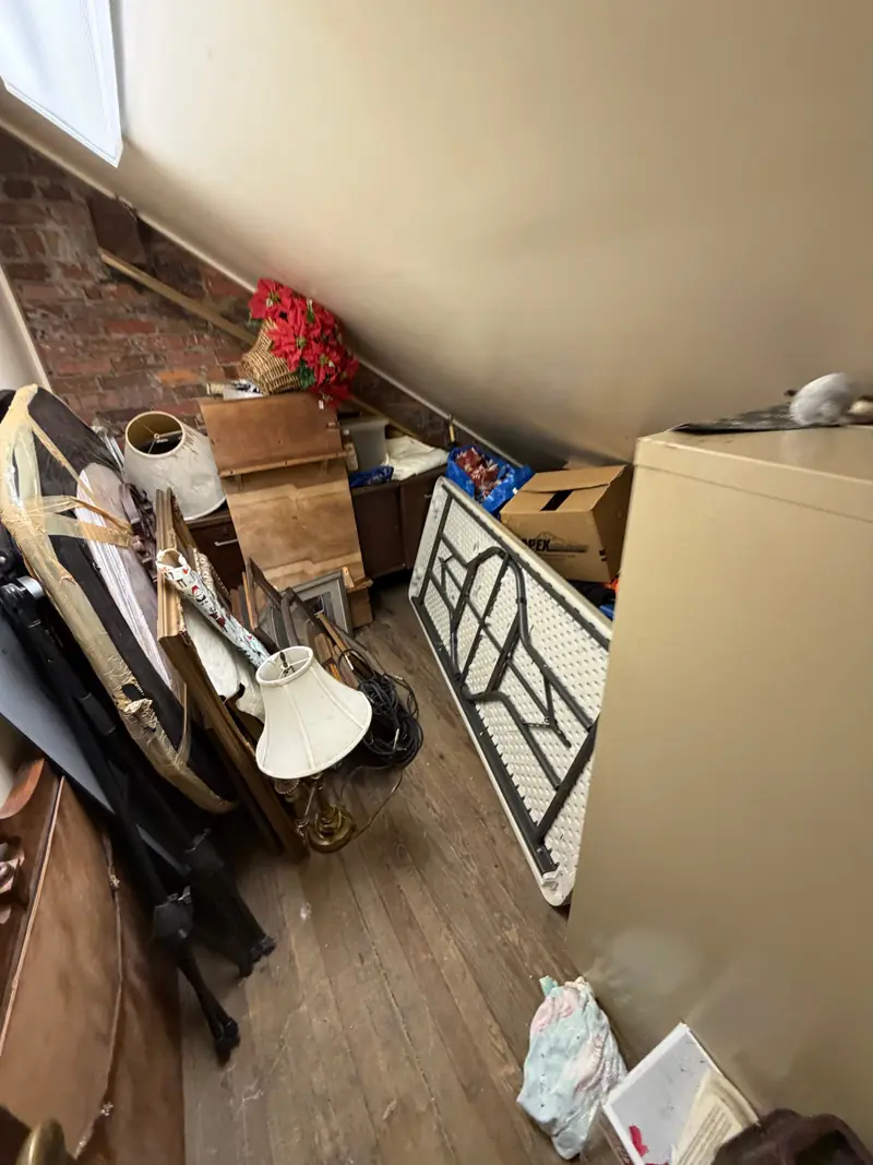 An attic room with a brick wall, crowded with lamps, a folding table, a filing cabinet and boxes.