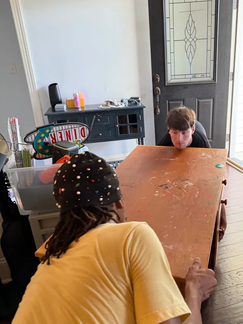 Two movers carrying a wooden dresser through a front room toward the open front door.