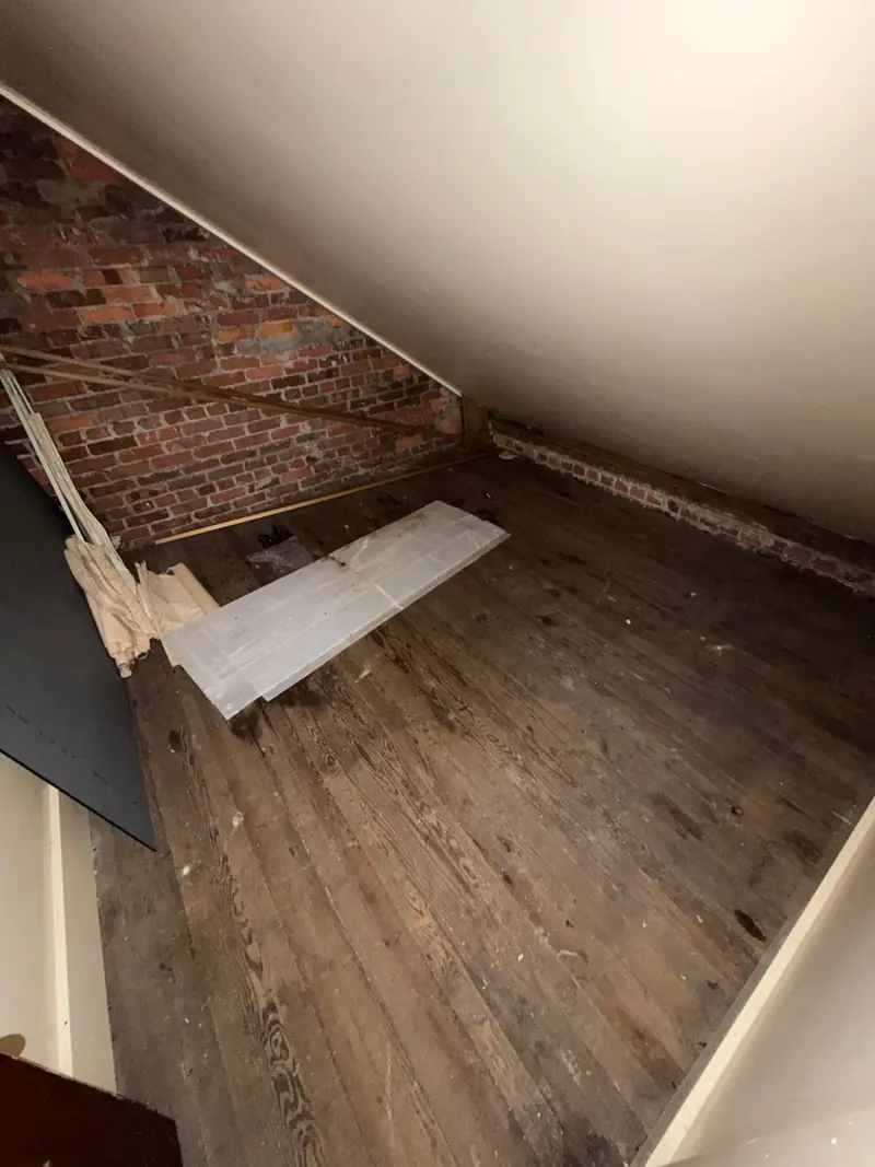An empty attic room with a sloped ceiling, an old brick wall and bare wooden floorboards.