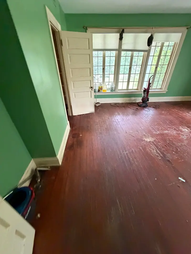 A bare room with green walls, tall windows and hardwood floors, with an upright vacuum and cleaning bottles by the window.