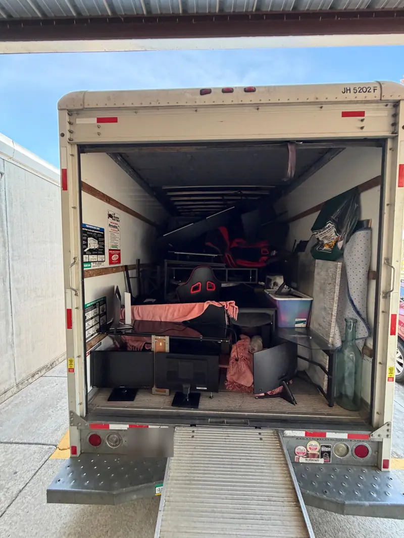 The open back of a rental truck loaded with chairs, desks, monitors and a rug at a storage facility.