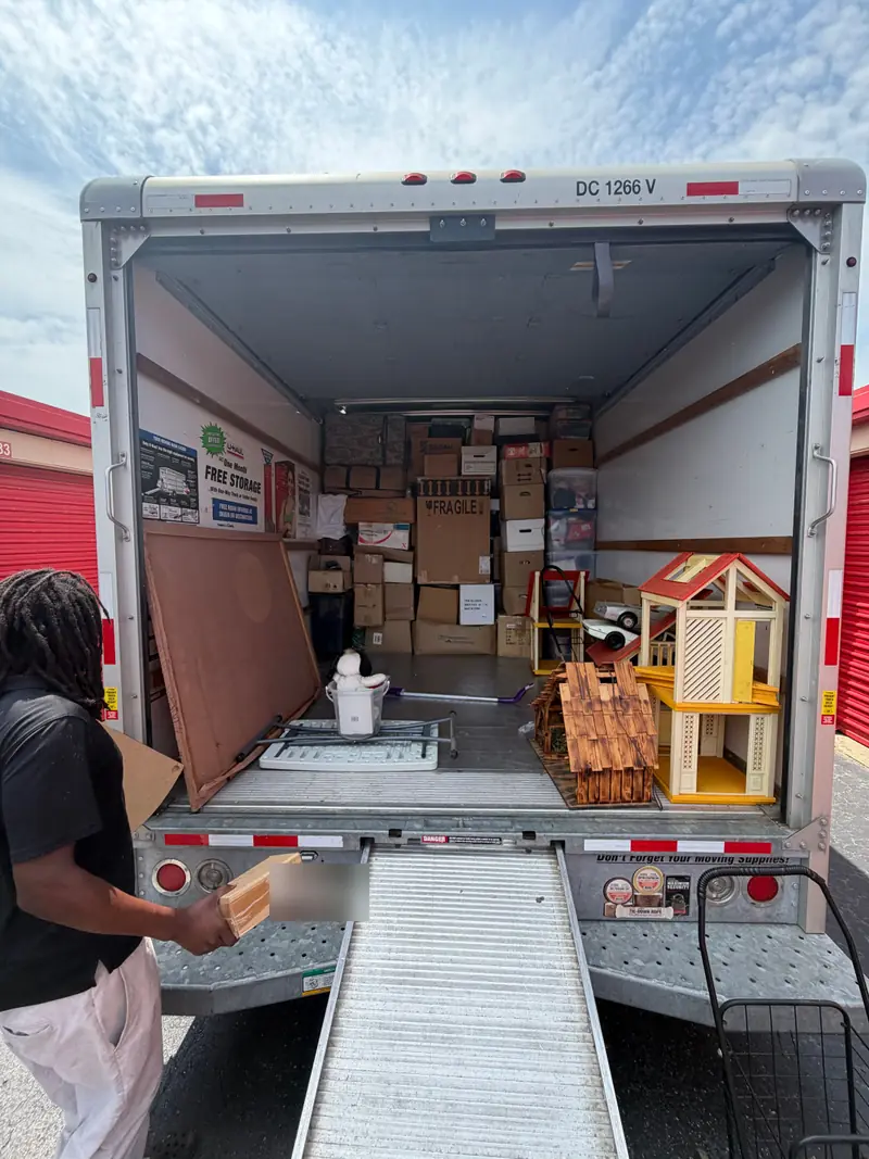 The open back of a loaded rental truck with its ramp down at a self-storage facility, and a mover carrying a box.